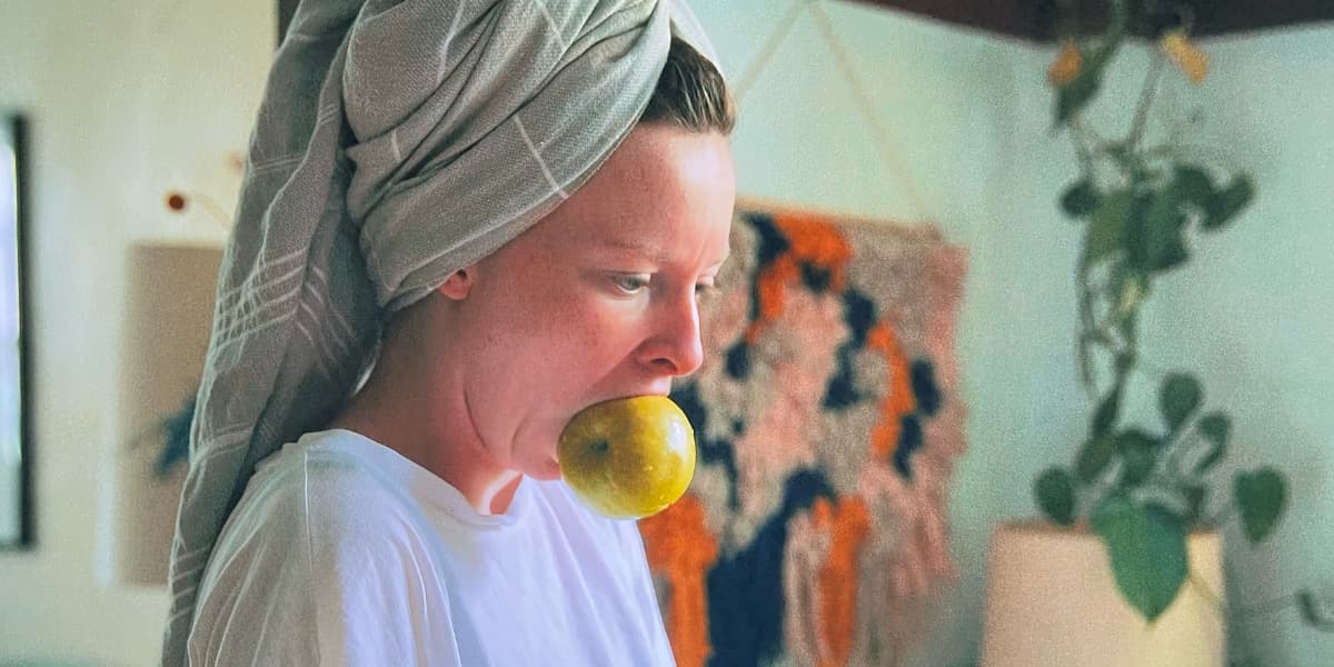 A woman with her hair in a towel and a green apple in her mouth looks worriedly down.