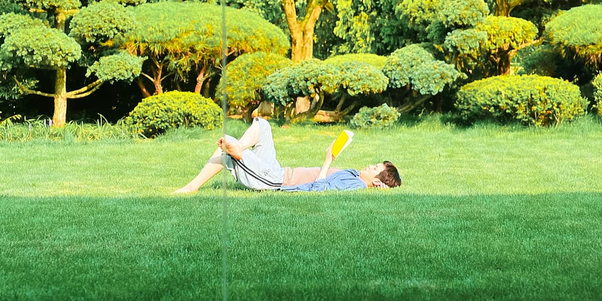 A boy likes on his back on the grass reading a book.