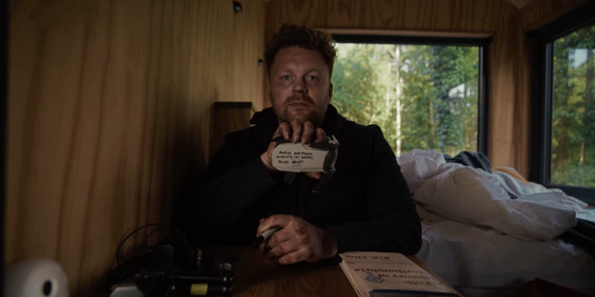A man shows a sign to the camera from his cabin in the woods.